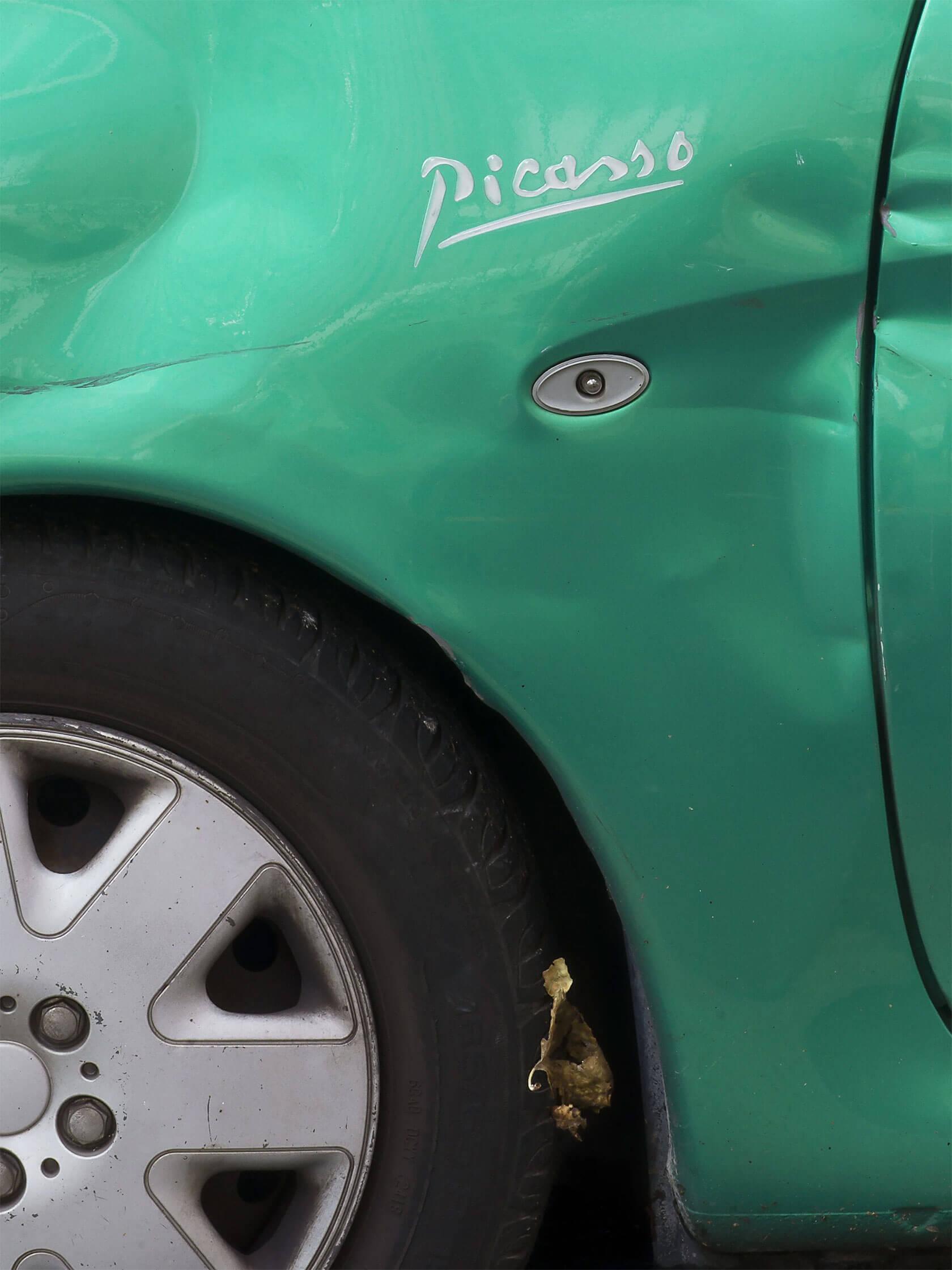 Close-up of a green Citroën Picasso showing its front wheel, scratched bodywork and silver Picasso badge.