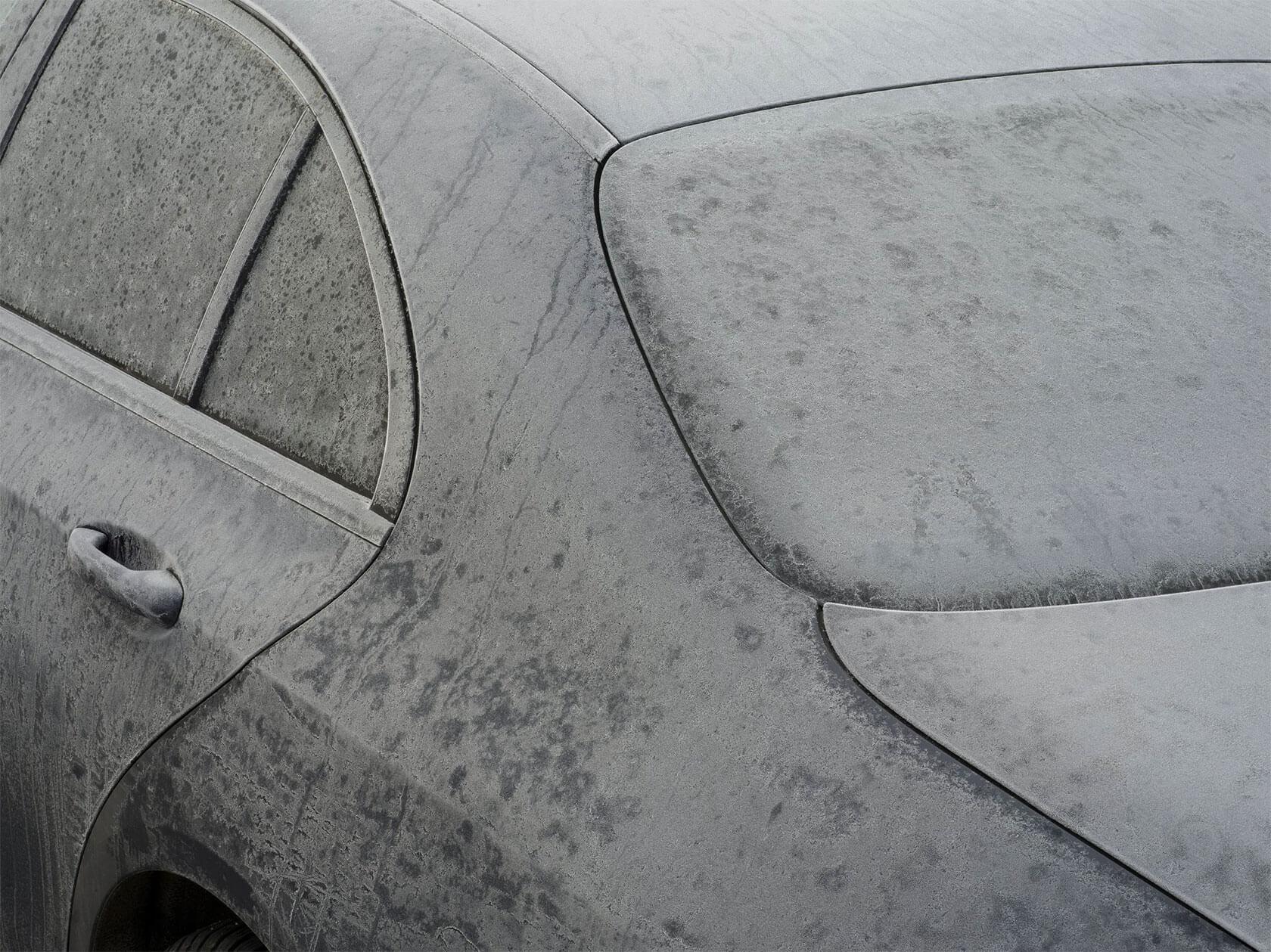 Close-up of a car coated in a thick layer of gray dust, highlighting the curved lines of its windows and bodywork.