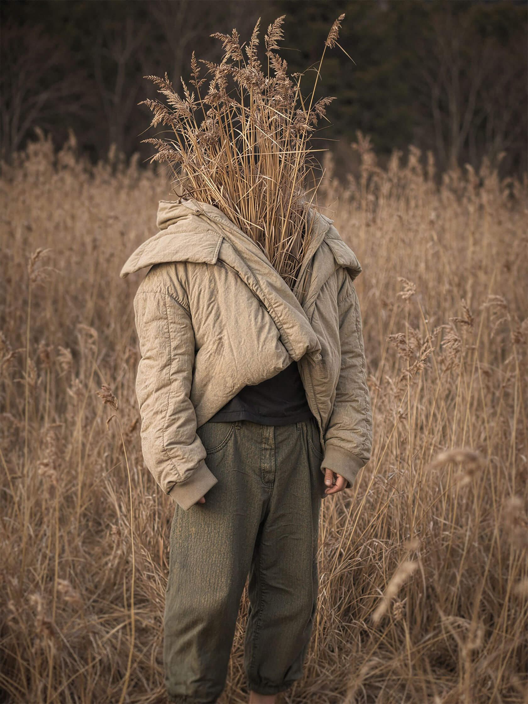 A surreal portrait of a person standing in a field of dry grass, with a bundle of tall reeds completely replacing their head.