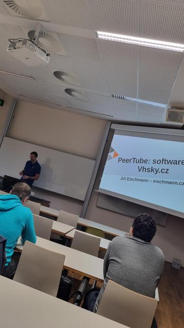 A white man in front of the screen with a peertube write on it.