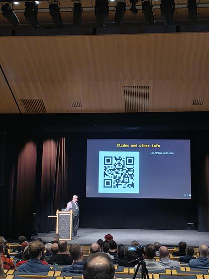 A man staying on a stage. A screen next to him shows a qr code.