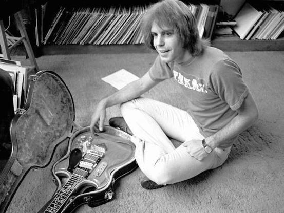 Bob Weir sits cross legged on floor with guitar in case, shelf of records behind him