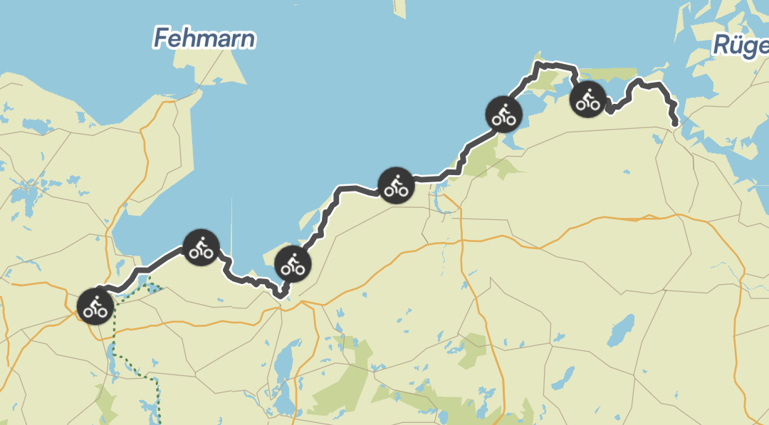 Eine Karte mit einer Radroute entlang der norddeutschen Küste. Der Weg ist durch eine dunkle Linie und mehrere Fahrrad-Icons markiert und führt von der Gegend um Lübeck in Richtung Stralsund. Die Inseln Fehmarn und Rügen sind in der blauen Ostsee zu sehen.