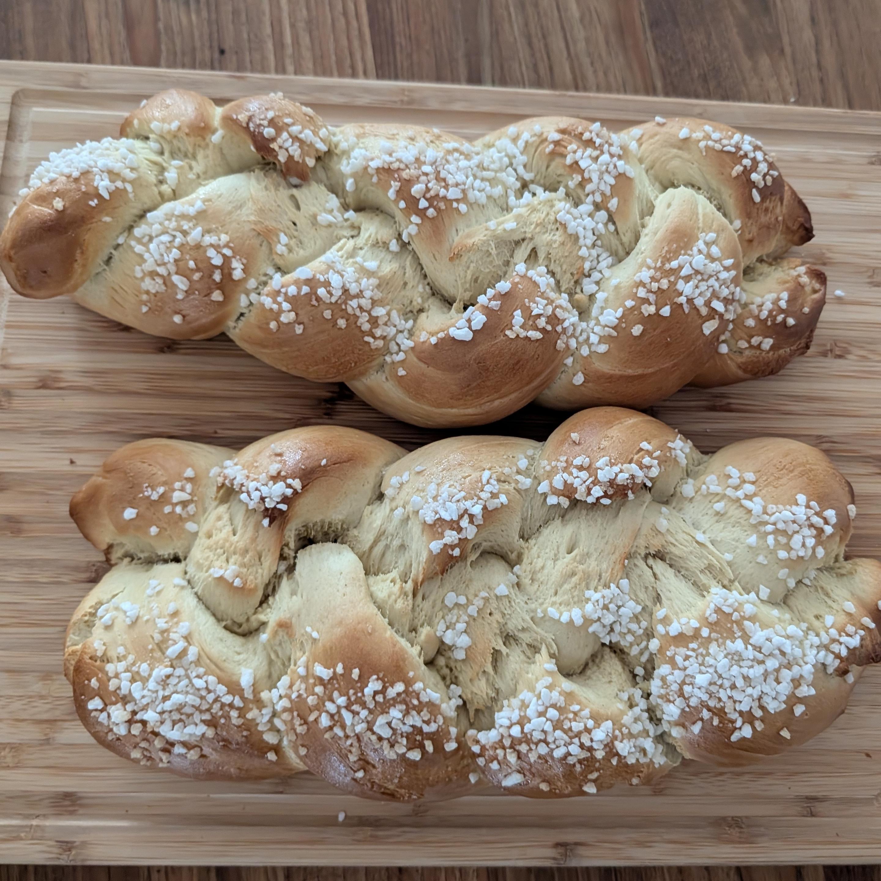 Zwei goldbraun gebackene Hefezöpfe liegen auf einem Holzschneidebrett. Die geflochtenen Brote sind üppig mit grobem weißem Hagelzucker bestreut. Die Textur zeigt weiche, faserige Schichten innerhalb der Flechtungen.