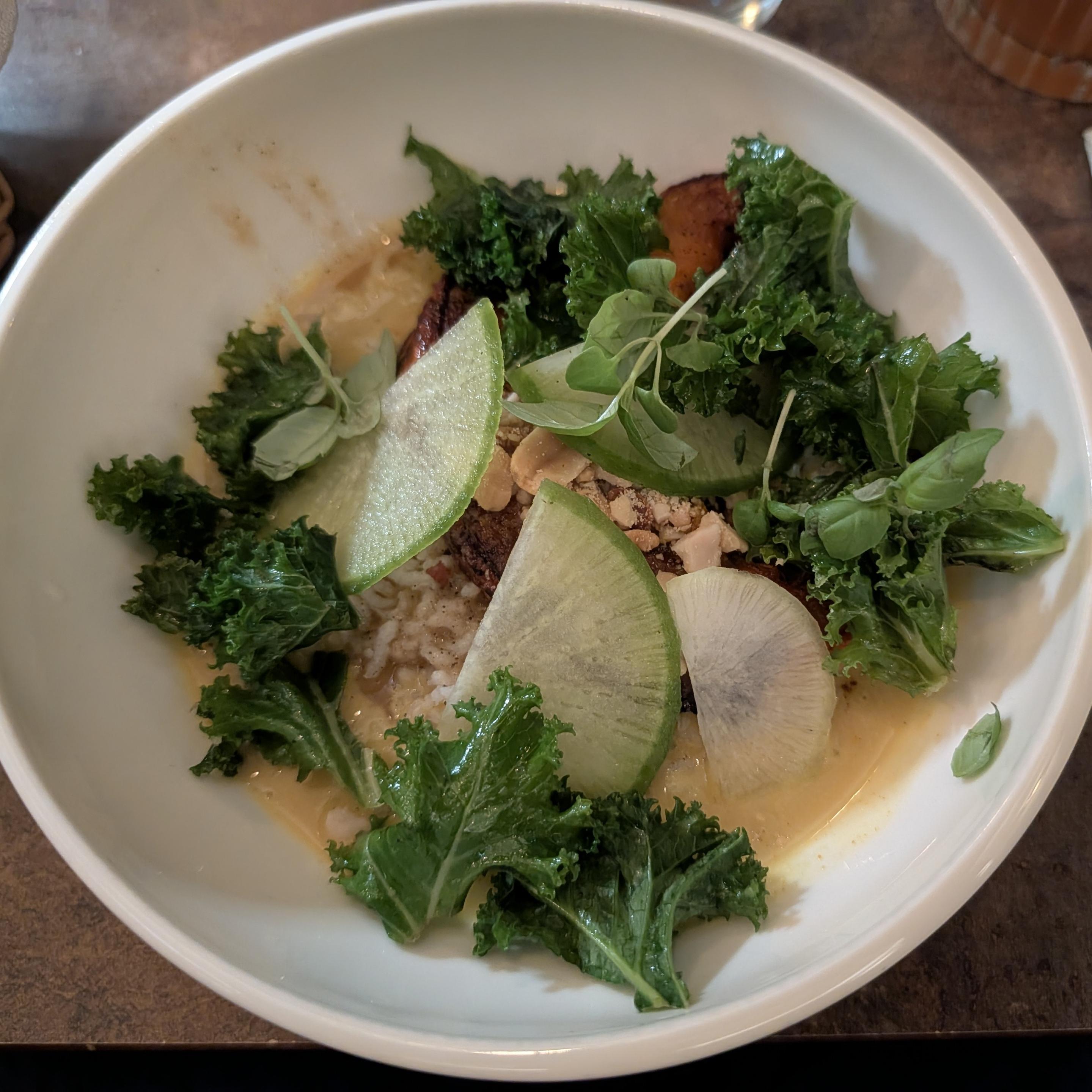 Eine weiße Schüssel mit einem cremigen Reisgericht, garniert mit kräftig grünem Grünkohl, dünnen Rettichscheiben und gerösteten Nüssen. Am Boden der Schüssel befindet sich eine helle Sauce.