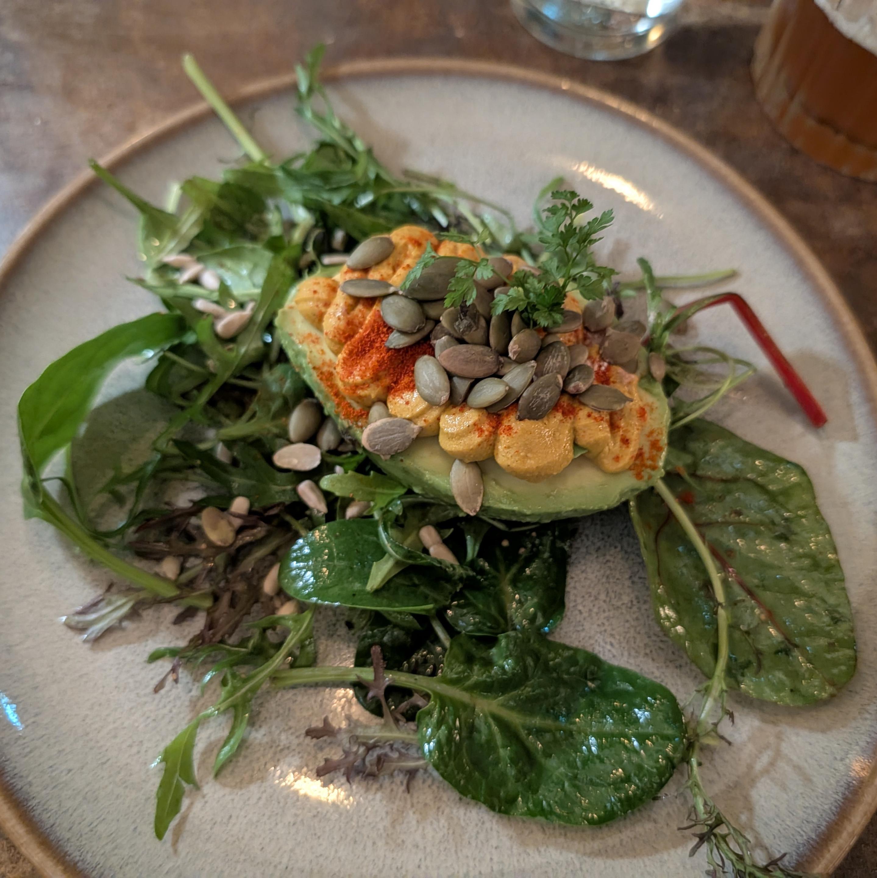 Eine halbe Avocado, gefüllt mit orangefarbenem Hummus, garniert mit Kürbiskernen und Paprikapulver. Sie liegt auf einem Bett aus frischem gemischtem Blattsalat auf einem gesprenkelten Keramikteller.