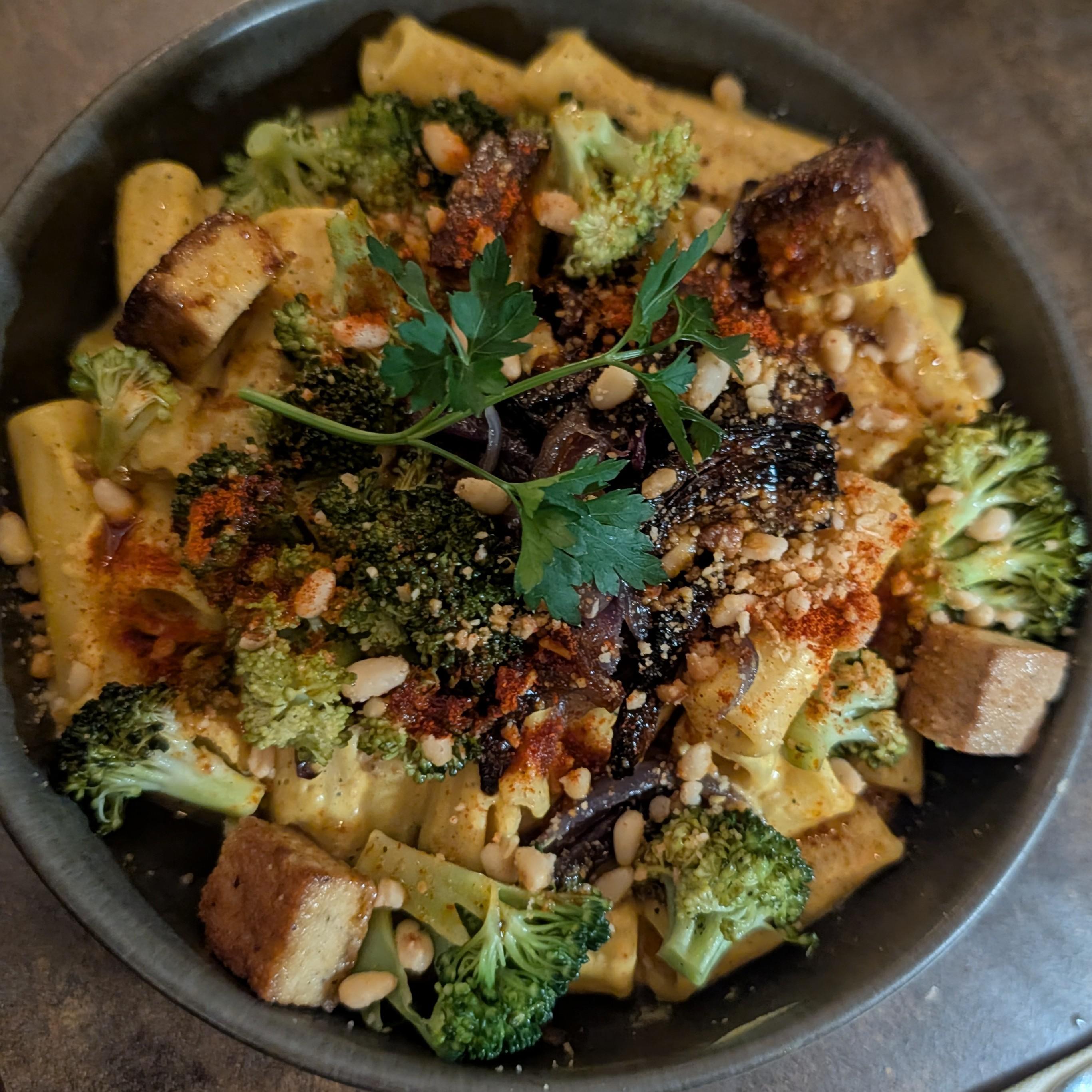 Eine herzhafte Nudelschale mit Rigatoni, gerösteten Brokkoliröschen, goldbraunen Tofuwürfeln und Pinienkernen. Das Gericht ist mit einem Zweig frischer Petersilie und einer Prise rotem Gewürz verfeinert.