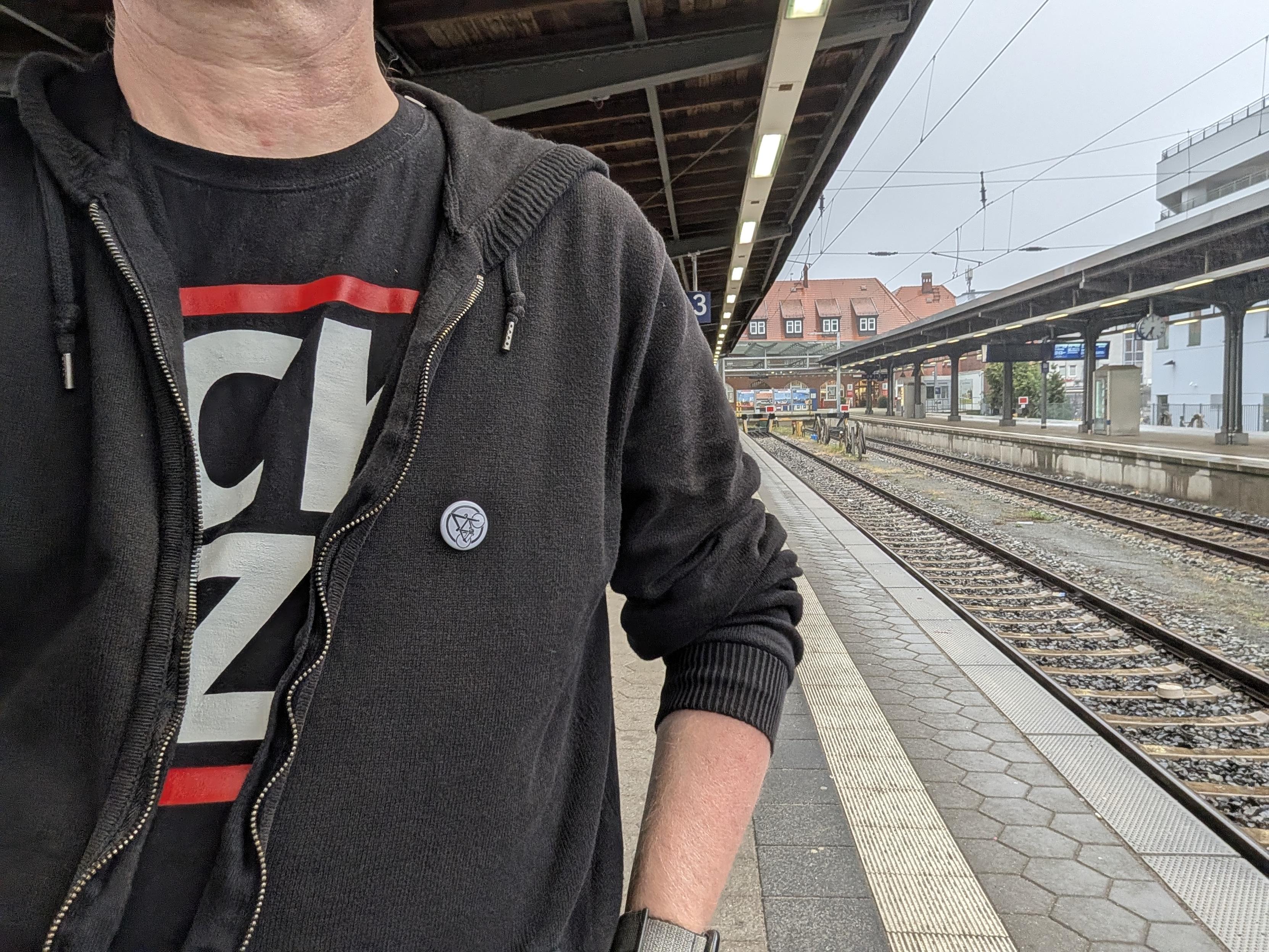 Eine Person steht auf einem Bahnsteig in Stralsund. Sie trägt eine schwarze Kapuzenjacke über einem T-Shirt mit großem Grafikaufdruck sowie einen kleinen runden Anstecker an der Jacke. Im Hintergrund erstrecken sich leere Gleise und Bahnsteigdächer unter bedecktem Himmel.