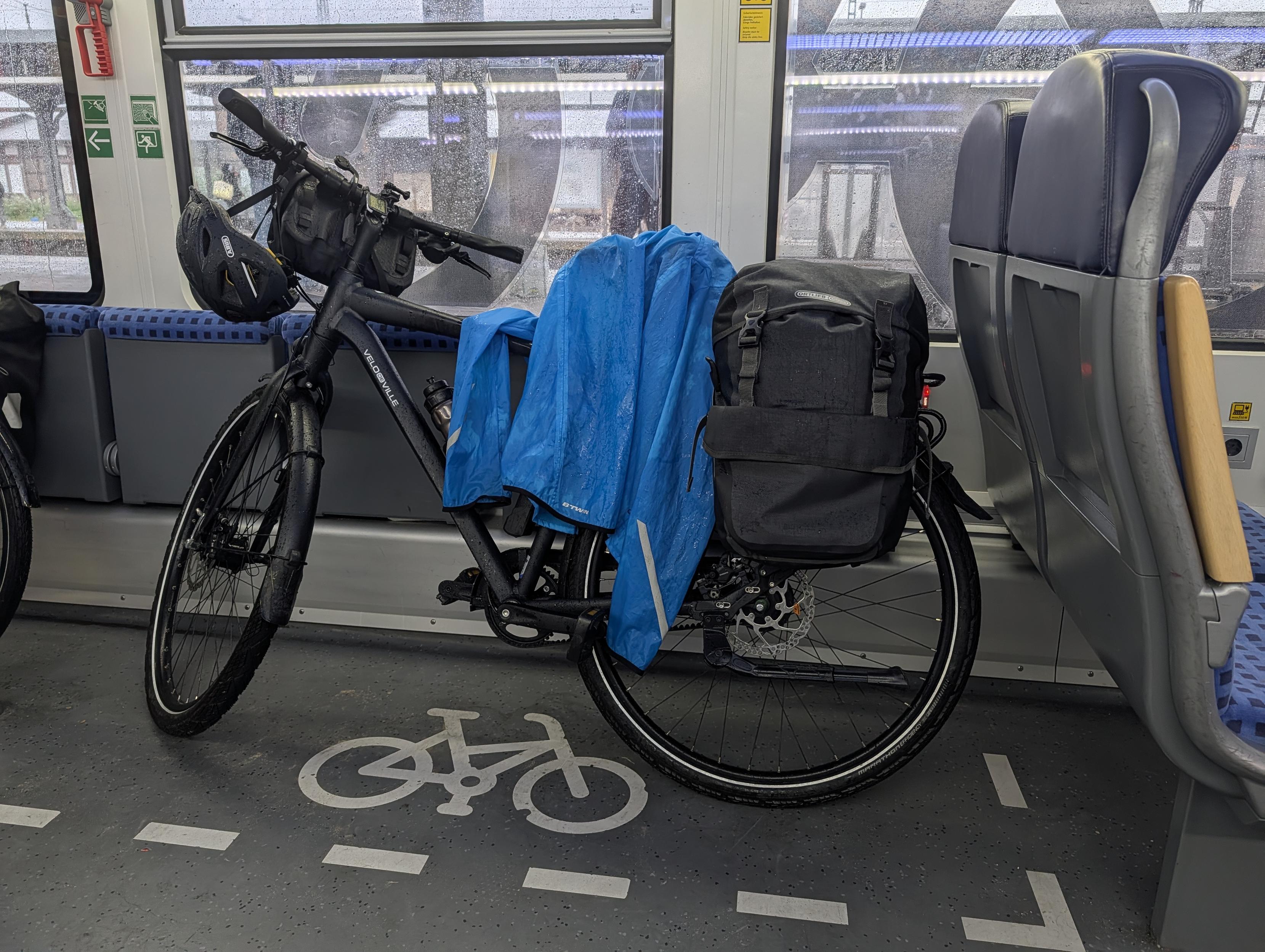 Ein nasses Tourenfahrrad steht im ausgewiesenen Fahrradbereich eines Zugabteils. Über dem Gepäckträger hängen eine leuchtend blaue Regenjacke und eine schwarze Packtasche, am Lenker ein Helm. Regentröpfchen bedecken das Zugfenster dahinter.