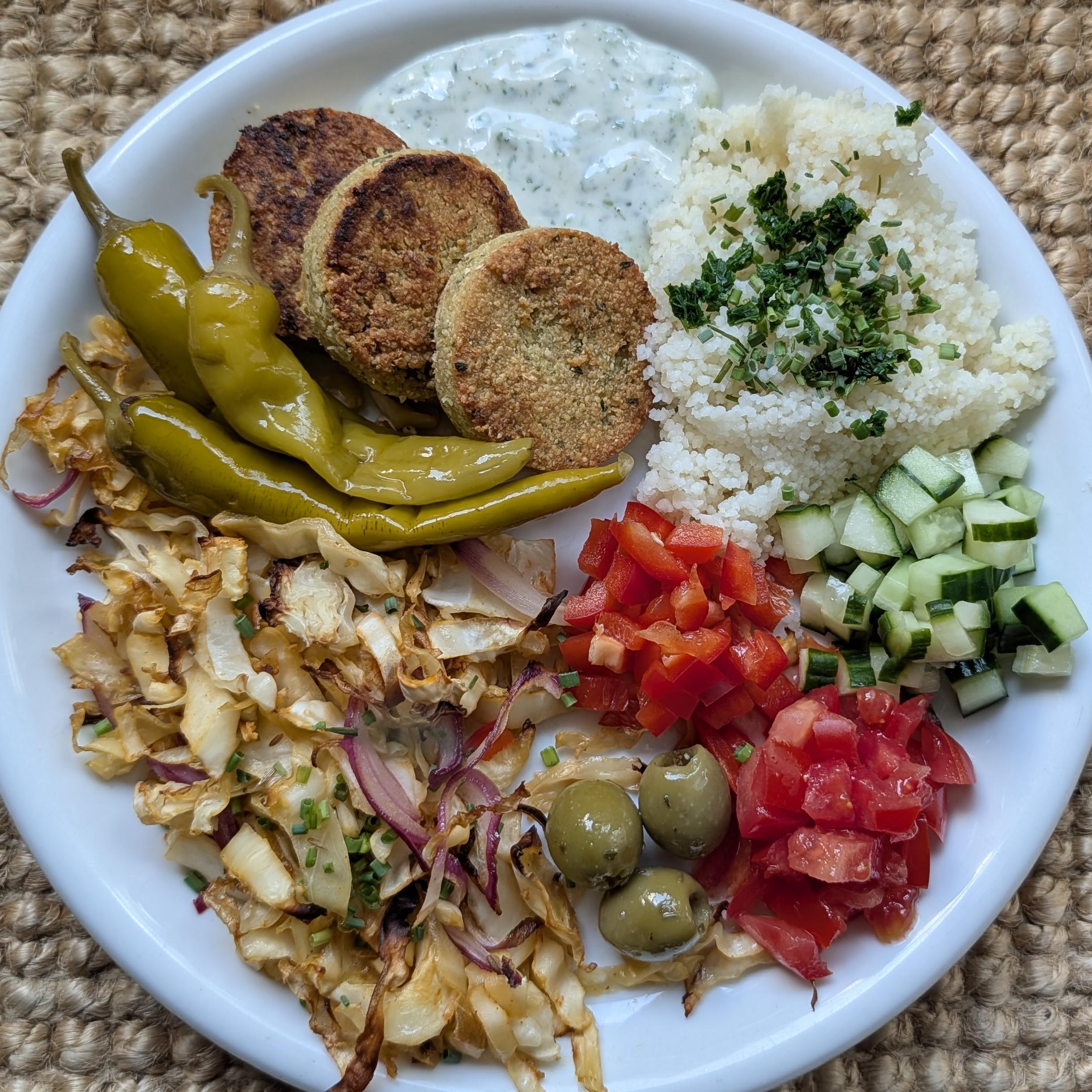 Ein bunt angemachter mediterraner Teller mit drei goldbraunen Falafel-Bällchen, Kräutercouscous, weißem Joghurtdip, eingelegten grünen Peperoni, grünen Oliven, gebratenem Kohl mit Zwiebeln sowie gewürfelten Gurken, roten Paprika und Tomaten.