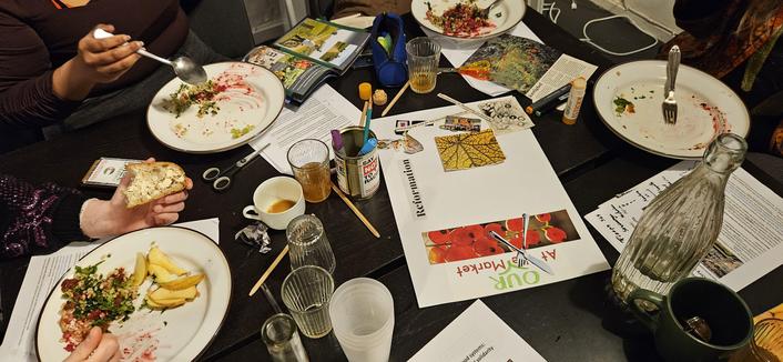 A messy table with food on plates, scissors, glasses with coffe and kombucha, glass bottle, open magazine for collage making, pencils, etc.