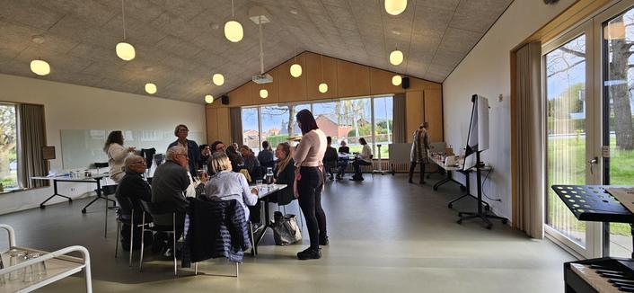 Several tables of people in a big room with many round lights hanging from the ceiling and large bright windows with green space outside. One person stands near a table talking.