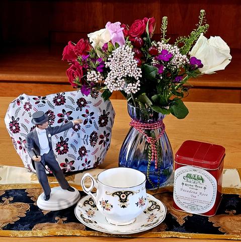 Tea tableau with a vase of colorful flowers seeming to burst into bloom at the command of a Fred Astaire statue in a dance pose with arm extended dramatically. There is vintage china, a floral tea cozy, an elegant quilted mat, and a red tin of Pasadena Rose black loose-leaf Tea to complement roses in the bouquet