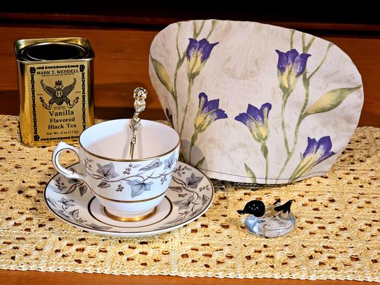 Tea tableau in soft yellow and parchment tones with purple accents. The cozy has subtle tulips, the vintage china has a muted ivy pattern, there is a small clear glass duck figurine with a black head and tail, the tea is Madagascar Vanilla black loose leaf from Mark T Wendell Teas. The base is a yellow crochet topper.