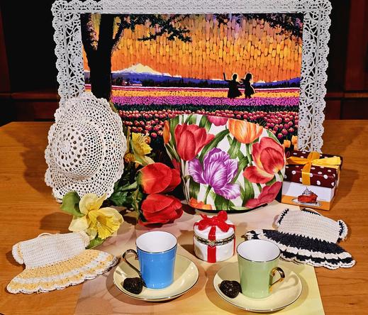 Tea tableau in front of a vibrant watercolor painting of two little girls in silhouette dancing in a field of tulips with Mt Baker in the background. The tea cozy is a tulip print, there are a lace hat and two crocheted doll dresses, a ceramic trinket box shaped like a present tied with a red bow, a gift box decorated with a birthday cake, a bouquet of tulips and daffodils, two vintage demitasse sets in sherbet colors, and two heart shapes of pressed tea leaves.