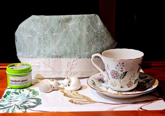 Tea tableau on an embroidered linen mat with two ceramic kitties curled up and sleeping, one pure white, the other half white and half gray. The cozy cover is a mint green and white sprigged print with white embroidered trim. The china is of a pleasing pattern of folk art florals in green, blue, and magenta on white. The tea is Mandarin Green loose leaf.