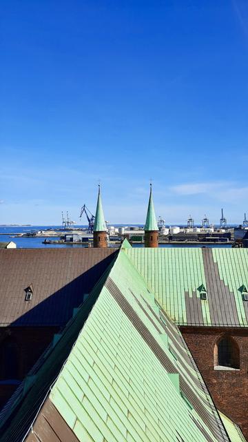 Inside the bell tower of Aarhus Domkirke, the mid-city cathedral in Aarhus, Denmark. We are 96 metres above the city, looking out through an unseen narrow window. We’re looking East towards the nearby harbour and open sea. Under a cloudless deep blue sky, we can see a glimpse of harbour buildings and industrial cranes and dark blue sea. On the turret of the old customs house in the distance, the Ukrainian flag is flying. The foreground of this upright image is filled by the cathedral’s main section below us and spires framing the harbour view. It is a redbrick cathedral with striking, bright copper-green roofs and spires.