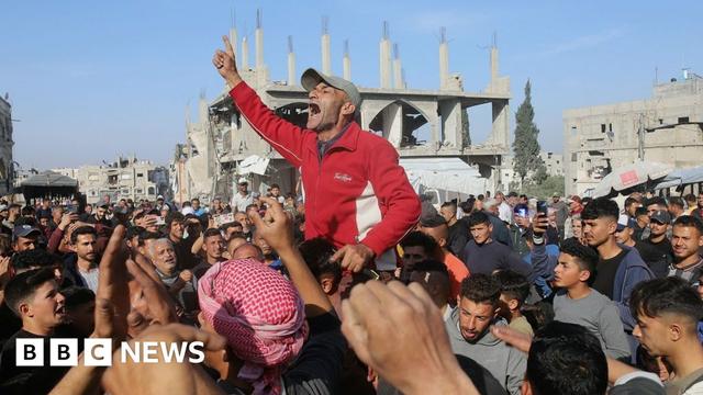A crowd of people line the street in Beit Lahiya, with a man in the centre sitting on another persons shoulders with a raised finger in the air as he shouts, with damaged buildings in the background against a blue sky.