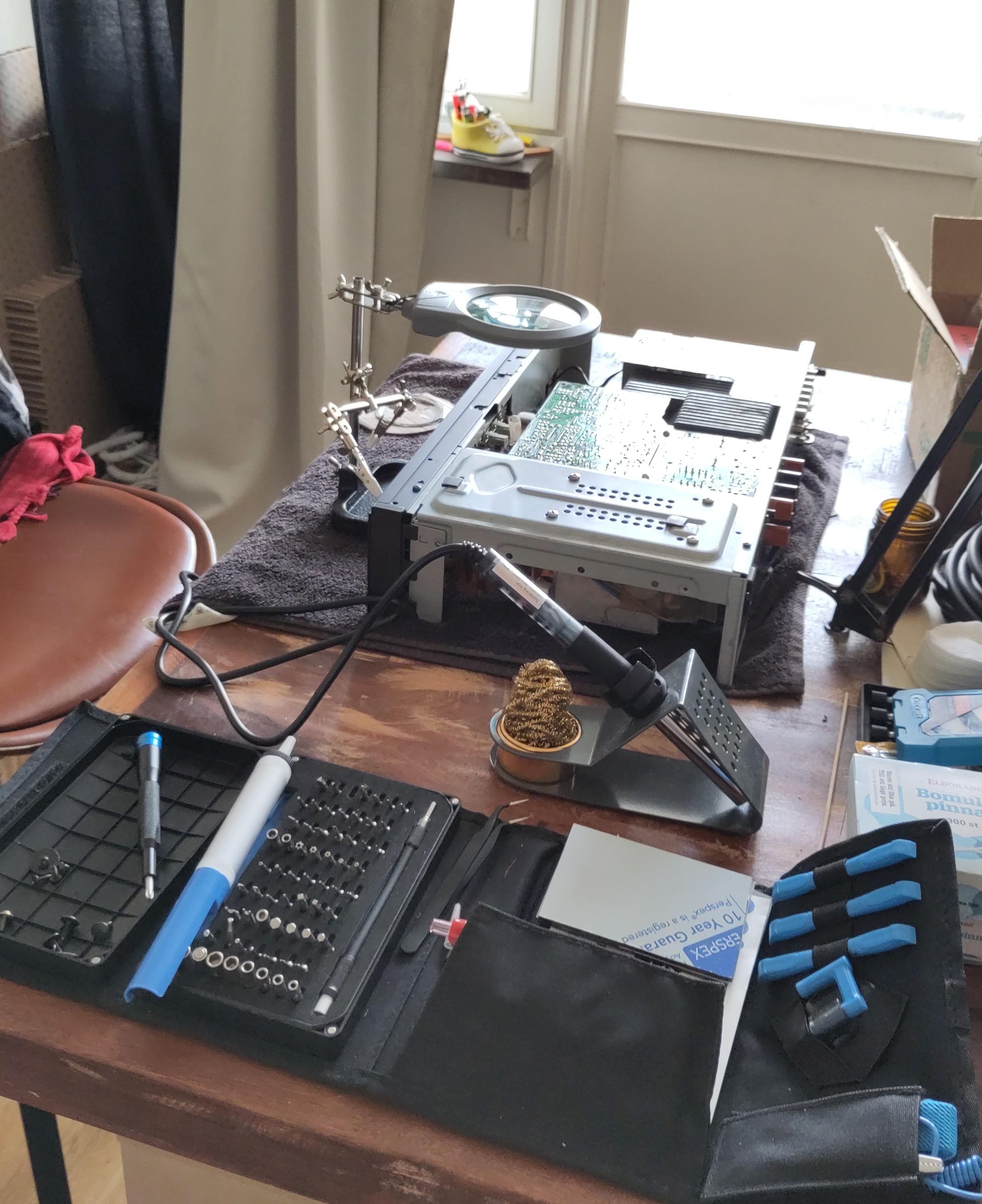 Arbejdsbord lagt op med loddekolbe, skruetrækkersæt, hjælpende hånd med lup, og en forstærker liggende på et håndklæde med åbent karosseri og bunden i vejret.