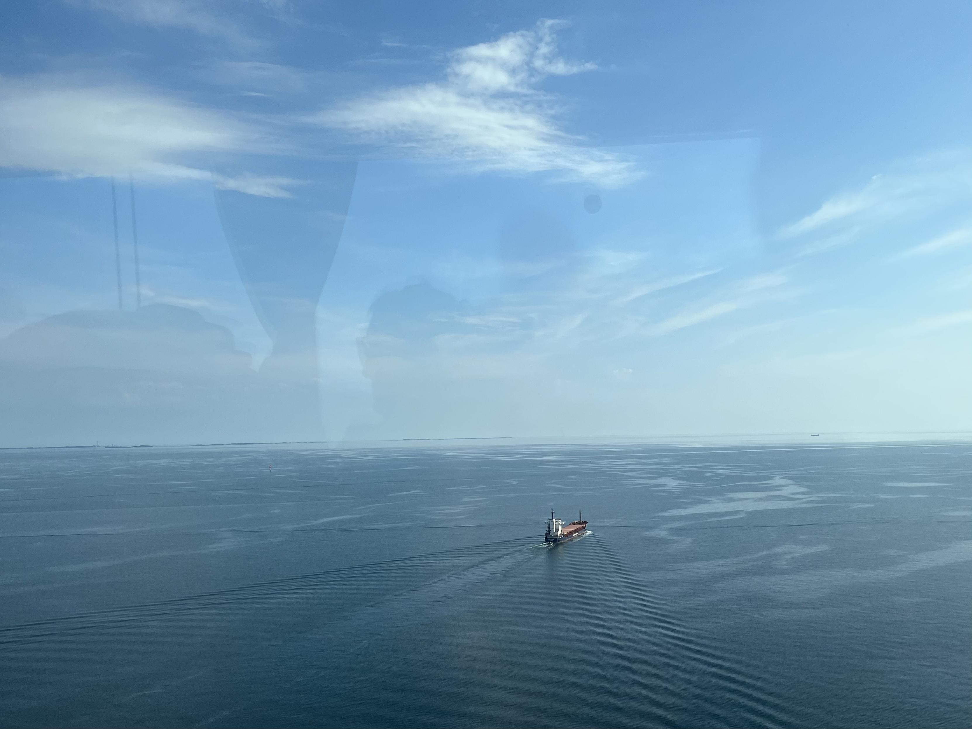 Foto ud af et busvindue af et fladt hav kun brudt af et skib i langsom sejlads, som trækker mildt kølvand og få bølger. Havet fortoner sig langt væk og bliver til lyseblå himmel, som strækker sig opad og bliver mørkere og får flere tottede skyer på.