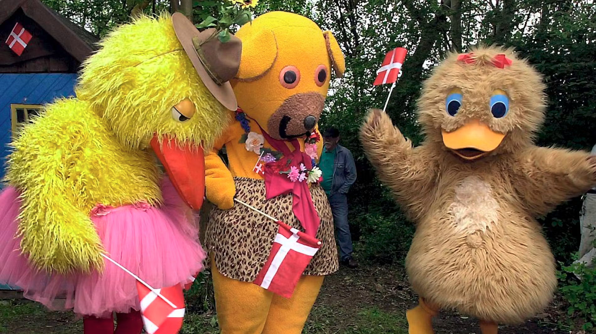 Figurerne Kylling, Bamse og Ælling fra børne-tv fjernsyn for dig Bamses Billedbog. Kylling har lyserødt balletskørt på, og hat, og holder et flag i hånden. Bamse har sine leopardprikkede badebukser og en blomsterkrans på, og holder også et flag, og Ælling har blomster i håret og holder et flag.