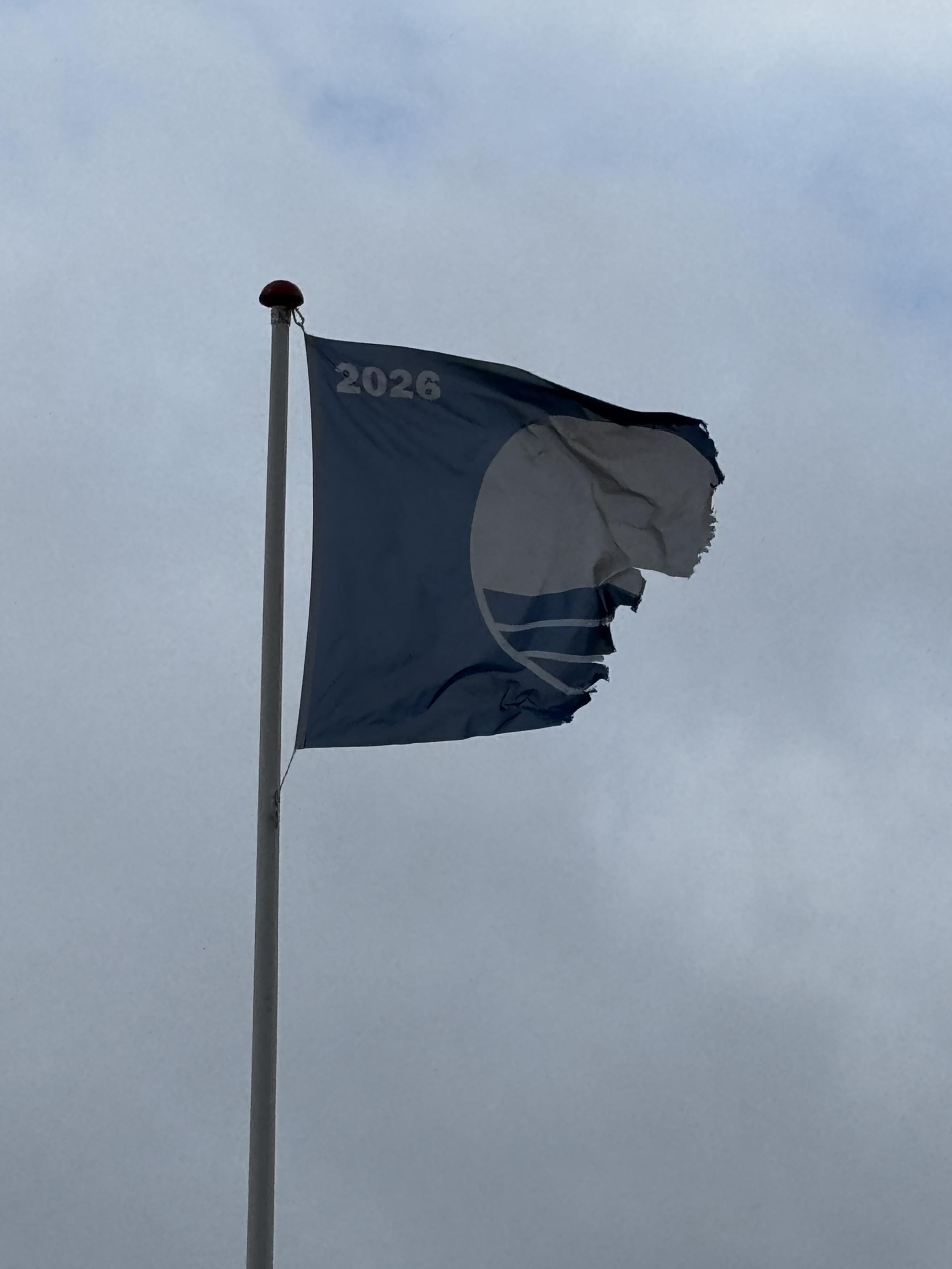Badeflag med årstallet 2026, udstrakt af en stiv kuling mod en grå himmel. Halvdelen af flaget mangler og enderne er flossede, som om den manglende halvdel er blæst eller revet af.
