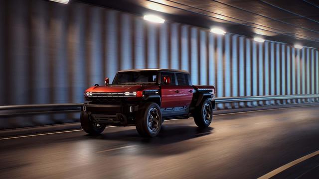 Red and black GMC Hummer EV pickup driving through a brightly lit highway tunnel, shown from a front three-quarter angle with motion blur in the background.