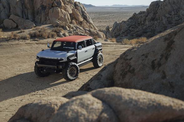 White GMC Hummer EV with an orange roof parked on a sandy desert trail among large boulders, shown from an elevated front three-quarter angle, with open desert and mountains visible in the distance.
