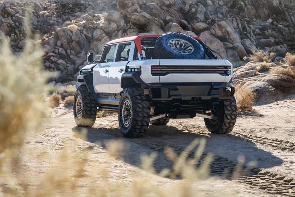 White GMC Hummer EV with a red roof parked on a sandy desert trail, viewed from the rear, with a full-size spare tire mounted in the bed, large off-road tires, and exposed suspension components, set against a backdrop of boulders.