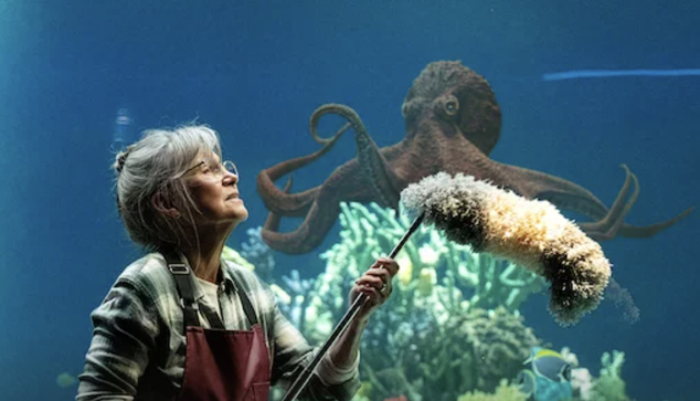 Actress Sally Field as Tova Sullivan, an older woman with gray hair in a bun and glasses, wearing a flannel shirt and apron and holding a long-handled brush, gazing up at Marcellus the giant Pacific octopus inside an aquarium tank. Coral and a small tropical fish are visible in the blue underwater background. Scene from the Netflix film Remarkably Bright Creatures.
