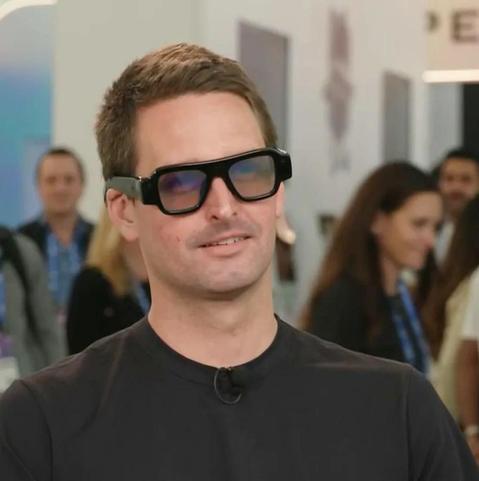 A man with short brown hair wearing thick-framed black Snap AR glasses and a dark T-shirt with a clip-on microphone, smiling during an interview. Blurred people and event signage fill the background.