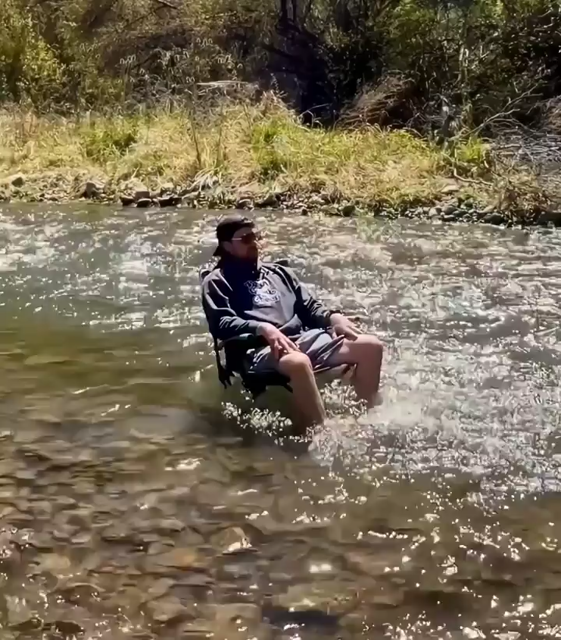 A man sitting on a chair in the middle of a creek with water flowing below him.