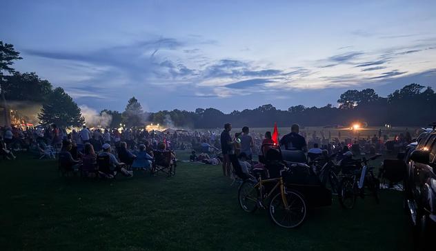 A large outdoor gathering at dusk with hundreds of people seated on grass and standing among bicycles, illuminated by gathering darkness and distant lights as the sky transitions from blue to twilight.