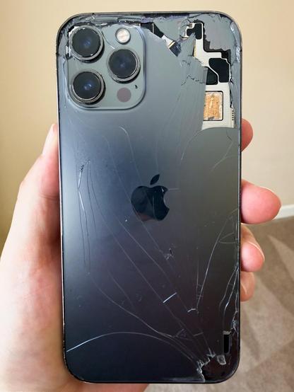 Damaged iPhone held in someone's hand, showing the back panel with severe cracks and damage, the camera array, and internal components including the SIM card tray and circuitry visible through an opening on the right side.