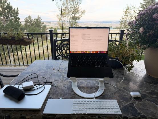 A laptop displaying code sits on a stone table outdoors, surrounded by a keyboard, trackpad, and charging cables, with a pastoral valley landscape visible beyond a decorative black railing.
