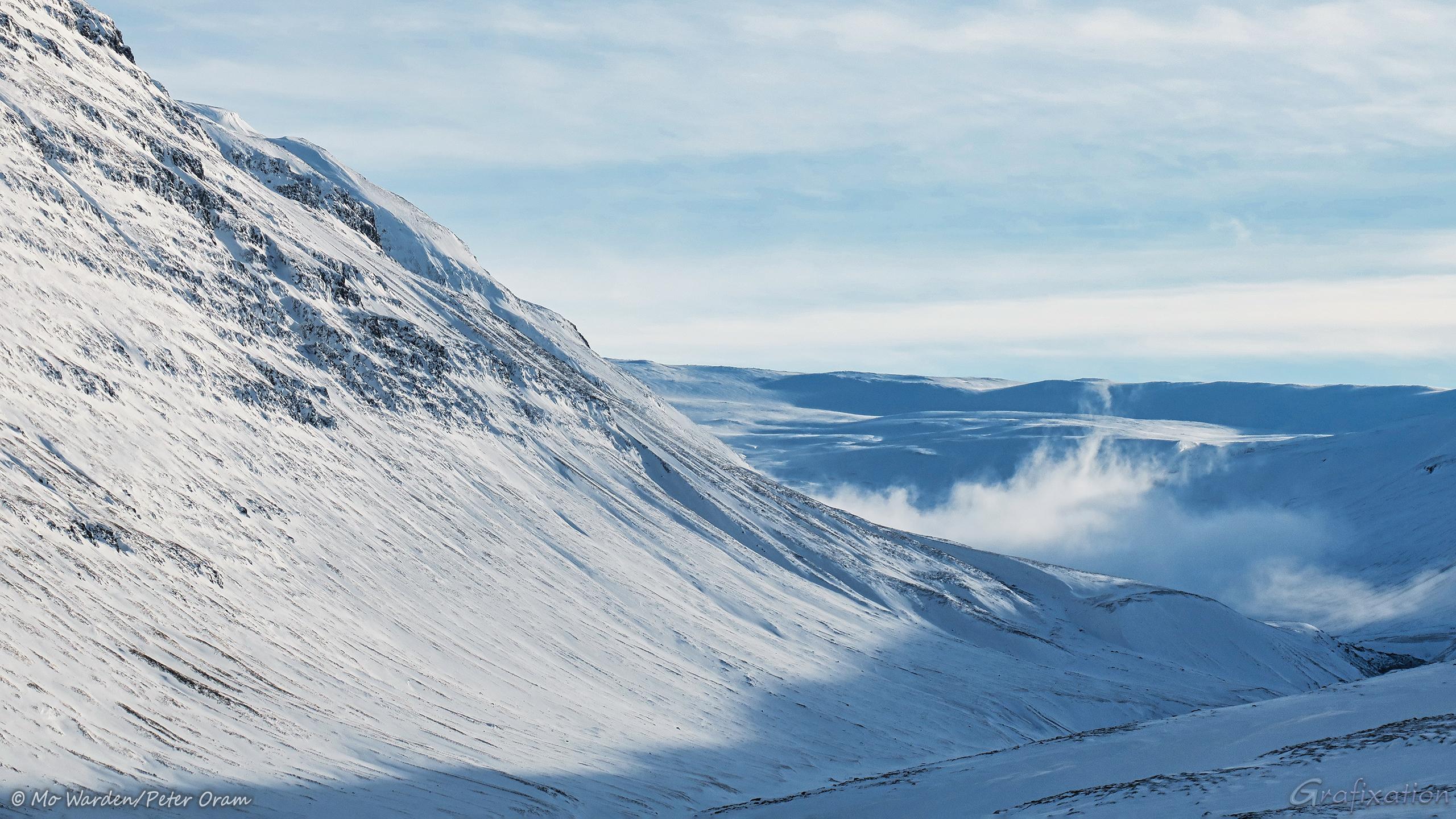 Another snowy mountain flank, this time with an unseen river running through it. The sky still cloudy but the valley is sunlit from the left. Dark rock strata are showing through the snow and ice. In a cleft of the valley, some wispy cloud is hanging, its top lit by the sun.