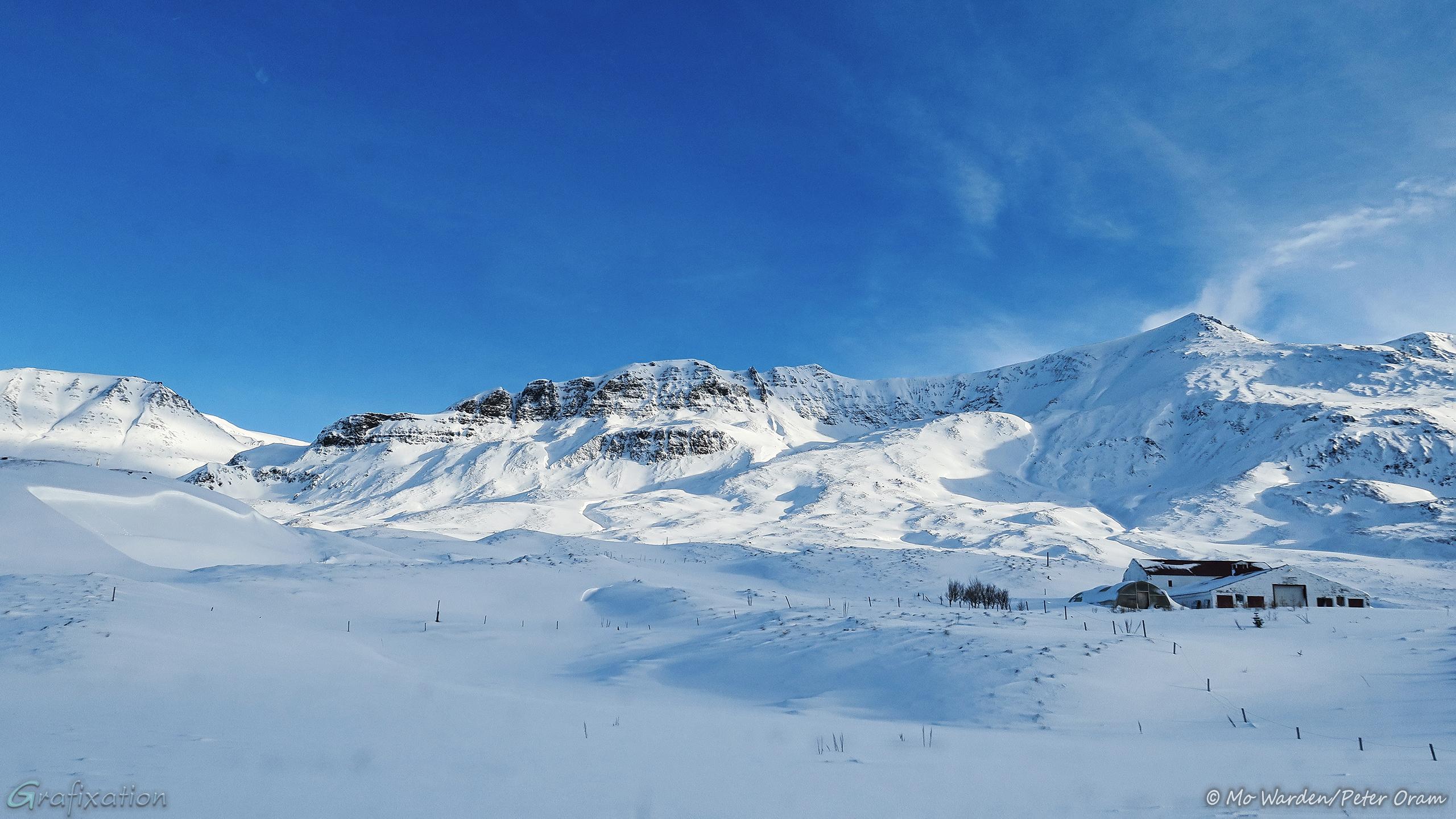 Another snowy mountain flank with the same intense blue sky above. The folds in the mountainside are accentuated by the light from the right. Some farm buildings are standing on the flank, lending scale. The foreground snow field is smooth and untrammelled, with fence posts half-buried in it.