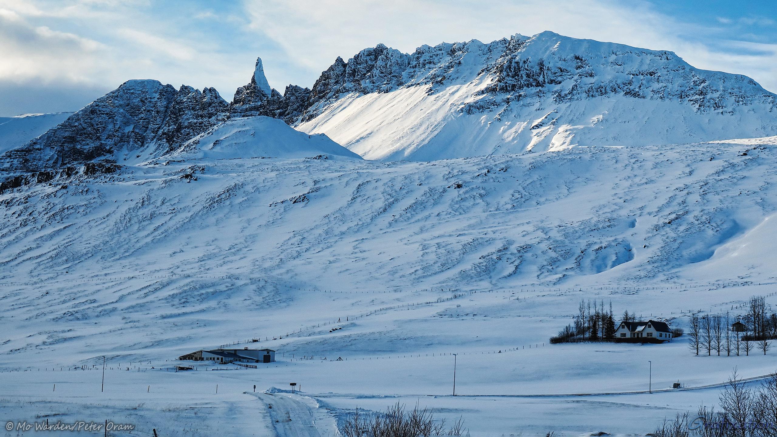 A mountain ridge with a distinctive conical centre peak, all lit from the left. The foreground is a road cleared through deep snow towards some farm buildings. There are bare trees scattered about, some in obviously planted lines. It's quite serene.