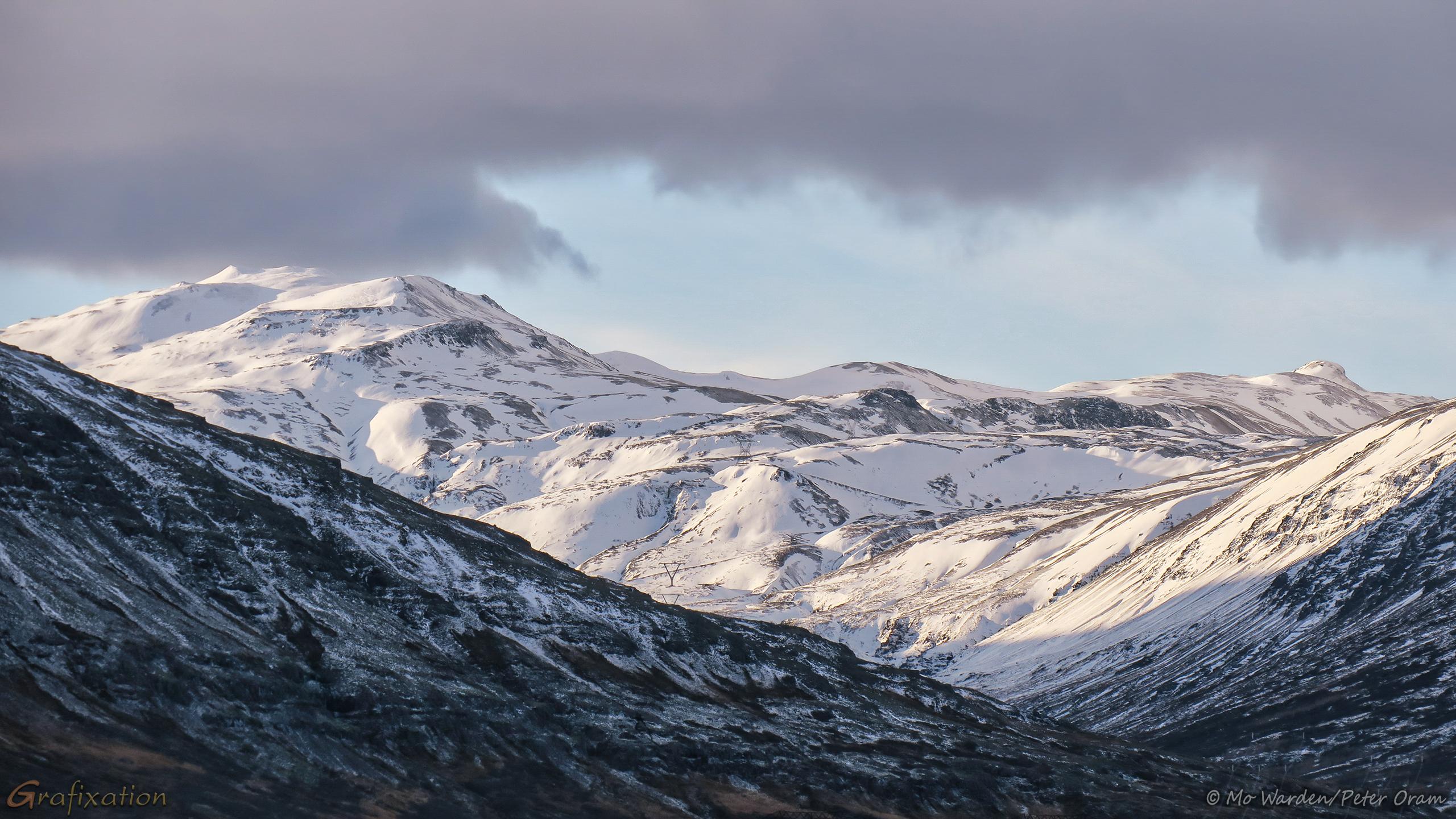 A snowy mountain with a very uneven surface, visible between two other peaks, the tops of which are not shown. The lumpy mountain is showing patches of dark rock amongst the snowfields. There is clear sky immediately above it, and a grey cloudbank above that.