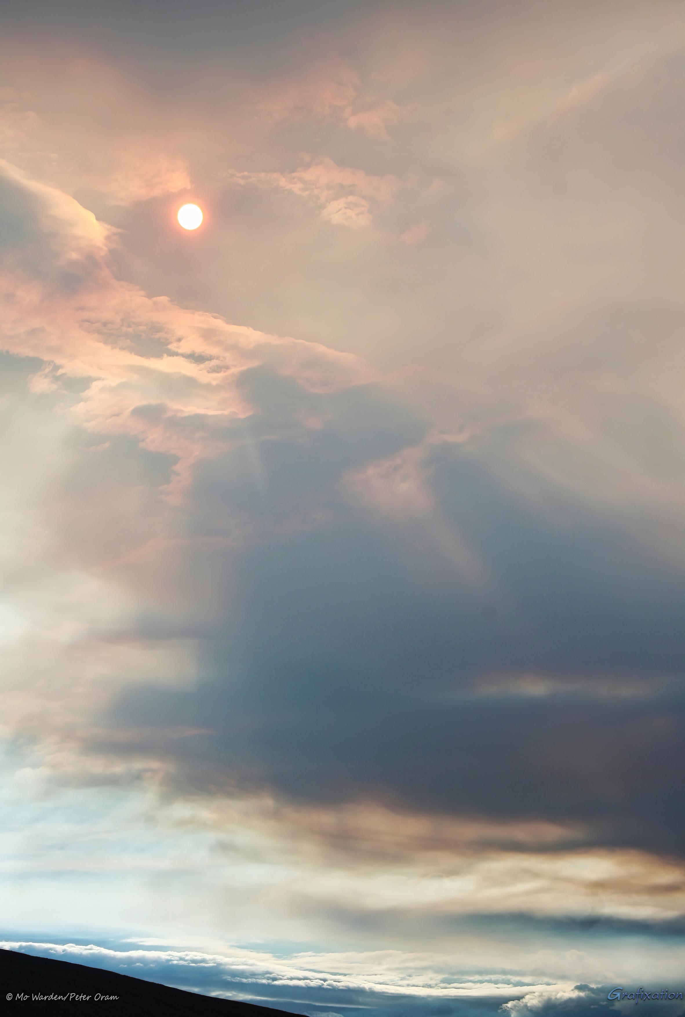 A colour photo of a sky filled with greyish orange clouds. There's a range of mountains at the bottom of the shot. The sun is shining through the cloud, producing a strange orange light. There are sunbeams across the clouds but even these are slightly unfamiliar.