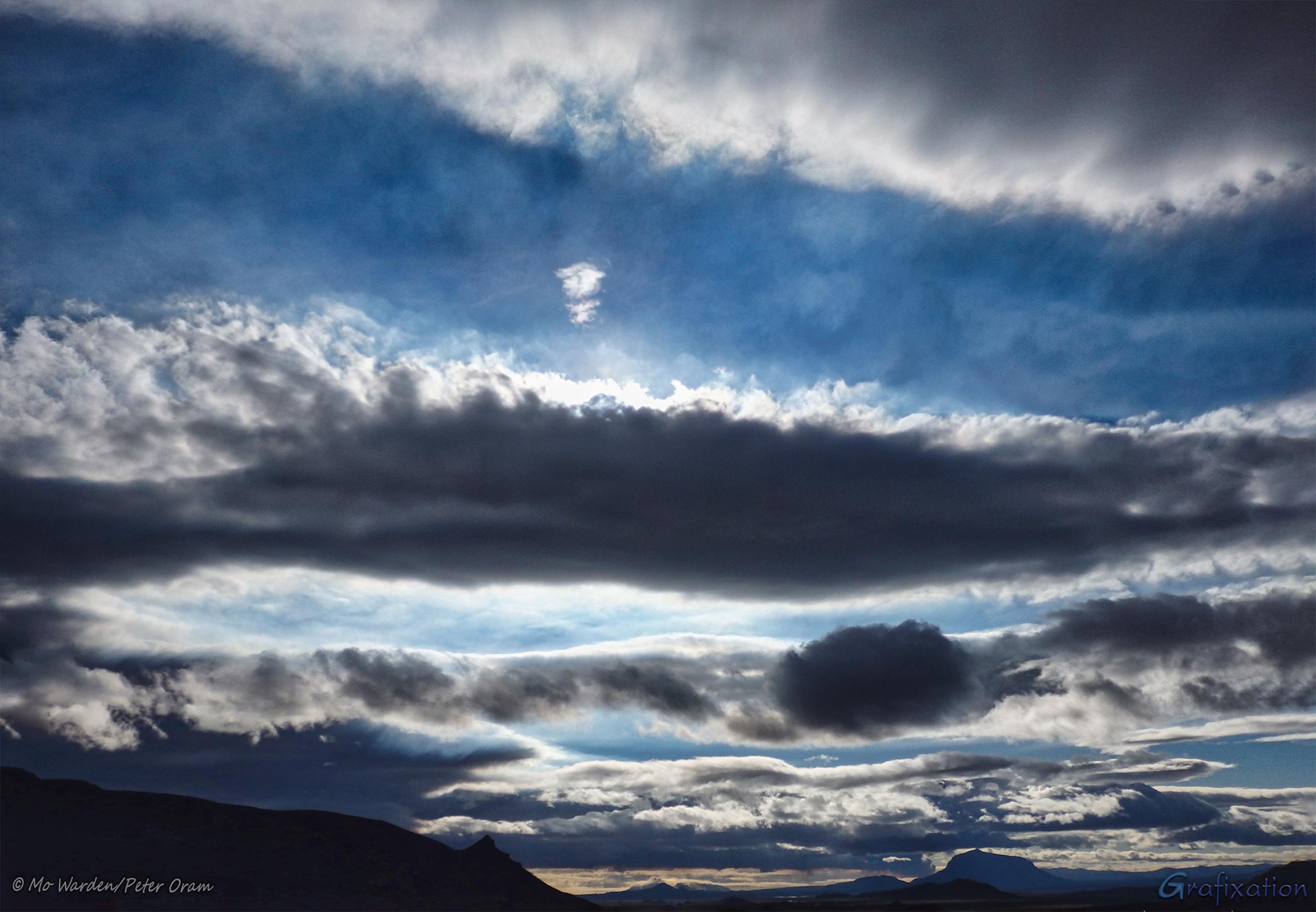 Another colour photo, this time looking away from the sun. The sky is filled with odd grey clouds, lit from above. Between them, a fine blue misty veil is visible against the clear sky. The base of the shot is a range of mountains, one of which is Herðubreið.