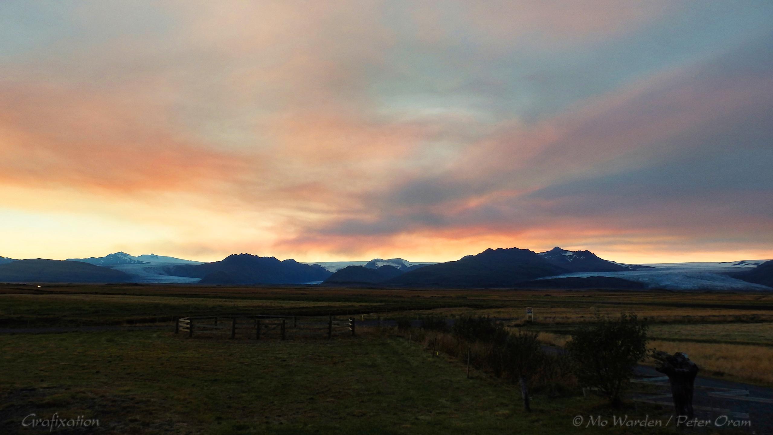 Sunset behind a mountain range with glaciers dropping to ground level. The foreground is indistinct brown pasture land. The sky is filled with darkening orange cloud with some lighter patches showing through which would usually, at this time of night, be darkening blue but instead are shades of yellow and grey. It's strangely beautiful.