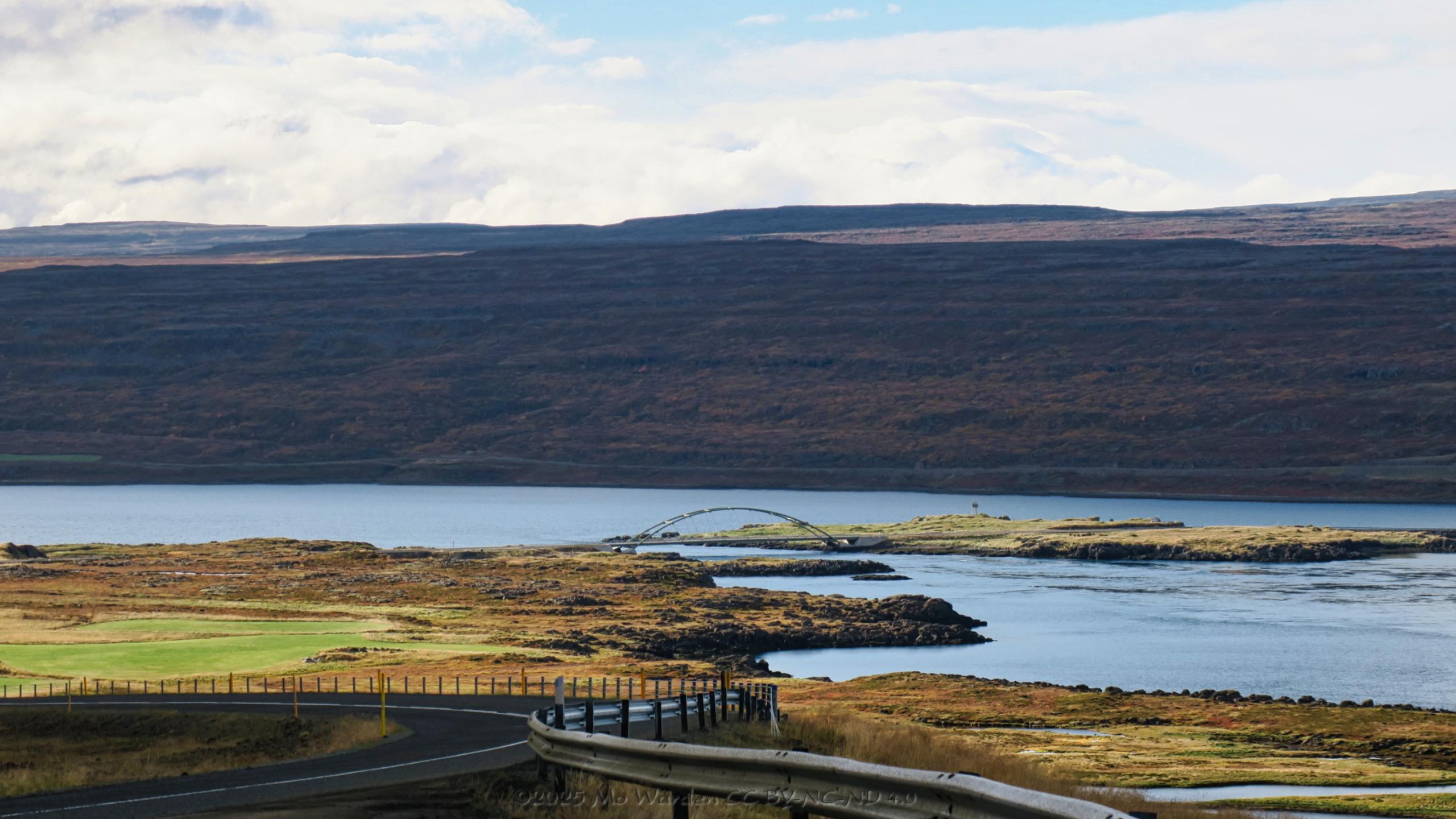 A colour photo of a landscape containing a body of water under a cloudy sky. The fjord and the browning vegetation on one bank is bathed in clear sunlight. Near the centre of the fjord is an island and a curved suspension bridge is connecting it with the road. The foreground is a steep curve of road with barriers along one side. In the distance is the shadowed flank of a mountain.