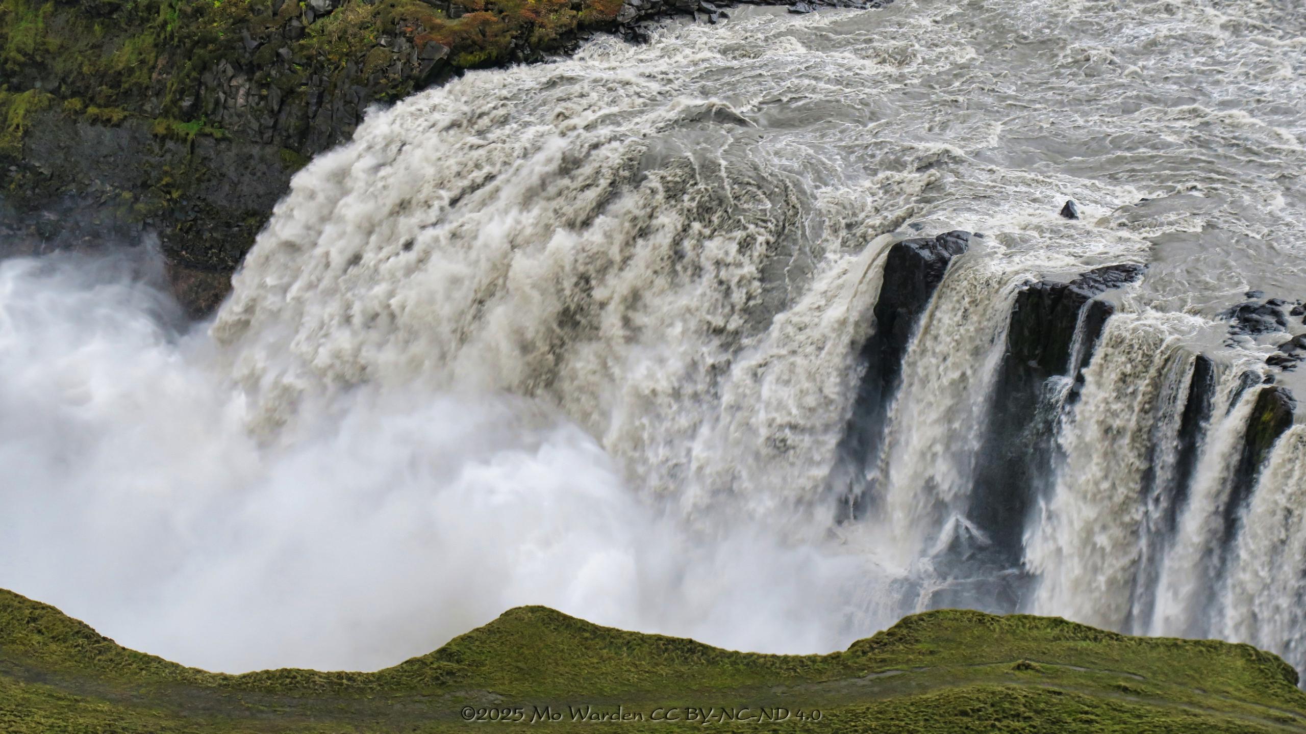 A colour photo of a waterfall plunging over a steep drop on a canyon floor. The water is white from turbulence but also from the particles being carried by it. Where the rockface is visible behind the water, it is a smoothed black surface. The foreground is the canyon edge and is covered in green mosses. The far wall is layered dark volcanic rock. There is a large amount of spray forming a dense mist at the waterfall's base.
