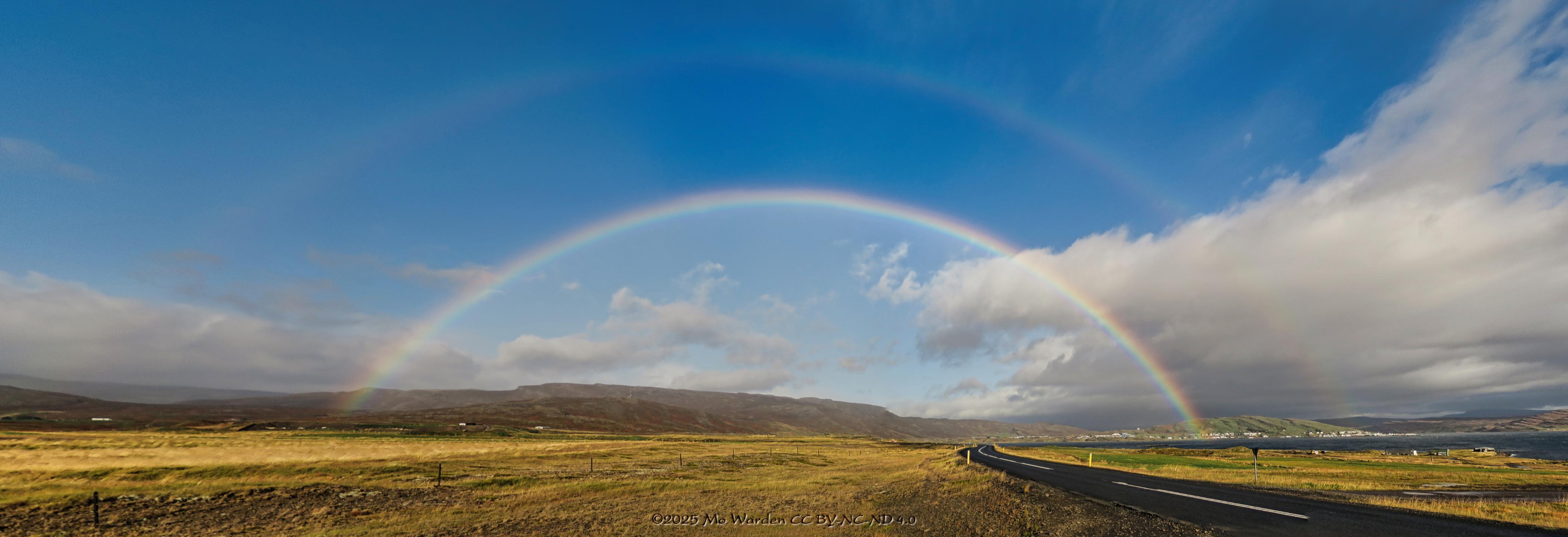A colour photo of a landscape under a blue sky with some low cloud. The foreground is golden grassland with fences dotted along it, and a black tarmac road snaking into the distance. On the right is a body of water and on its shore is a town. In the centre of the shot is the arc of a gorgeous double rainbow of about 140°, strong and clear, its faint companion just above it.