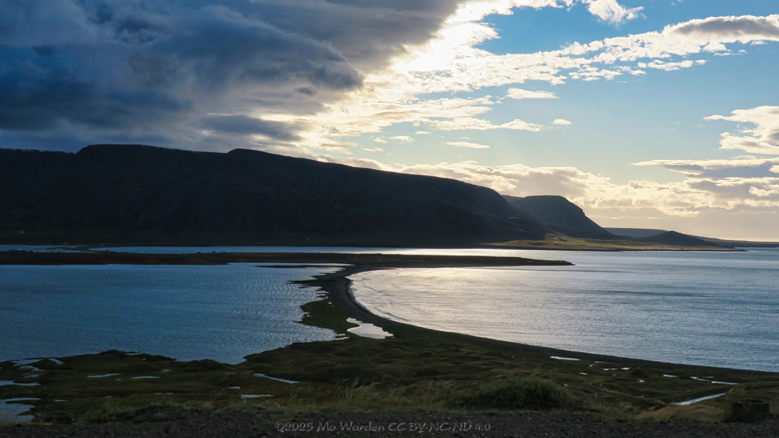 A colour photo of a body of water with a stretch of gravel and rock separating it into halves by connecting with a central promontory. The horizon is a silhouetted mountain range. The sky is cloudy on the left, and the solidity has a distinct edge which is directly above the isthmus. On the right, the sky is clearer.