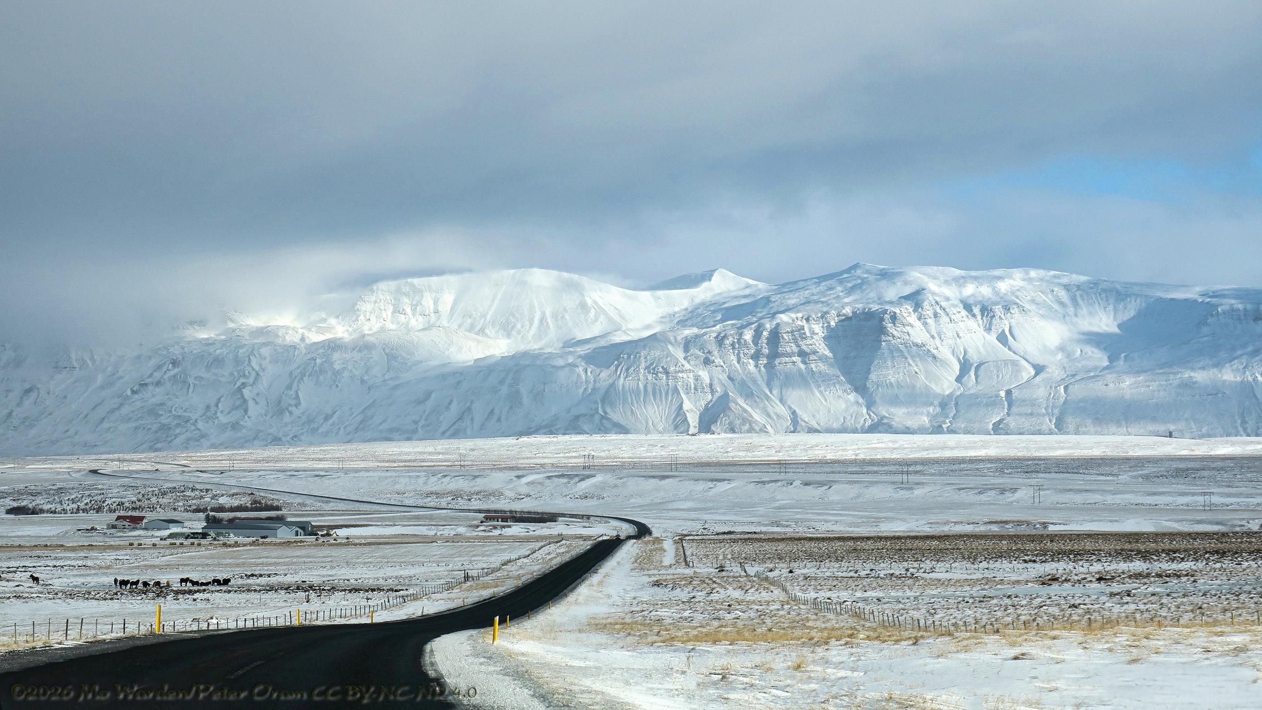 A colour photo of a mountainous landscape covered in snow and ice. The sunlight is from the right, and a heavy cloudbank is hanging just above the mountain tops, obscuring some of them. From the bottom left corner, heading towards the lower centre and then snaking away to centre left is a metalled road, clear black against the white snow. Yellow snow poles line its edge. On the horizon, the range of mountains is completely covered in snow and ice apart from ridges where the wind has scoured down to the dark rock. Winding gulleys of waterfalls can be seen on the right. On the left are farm buildings with horses in the field.