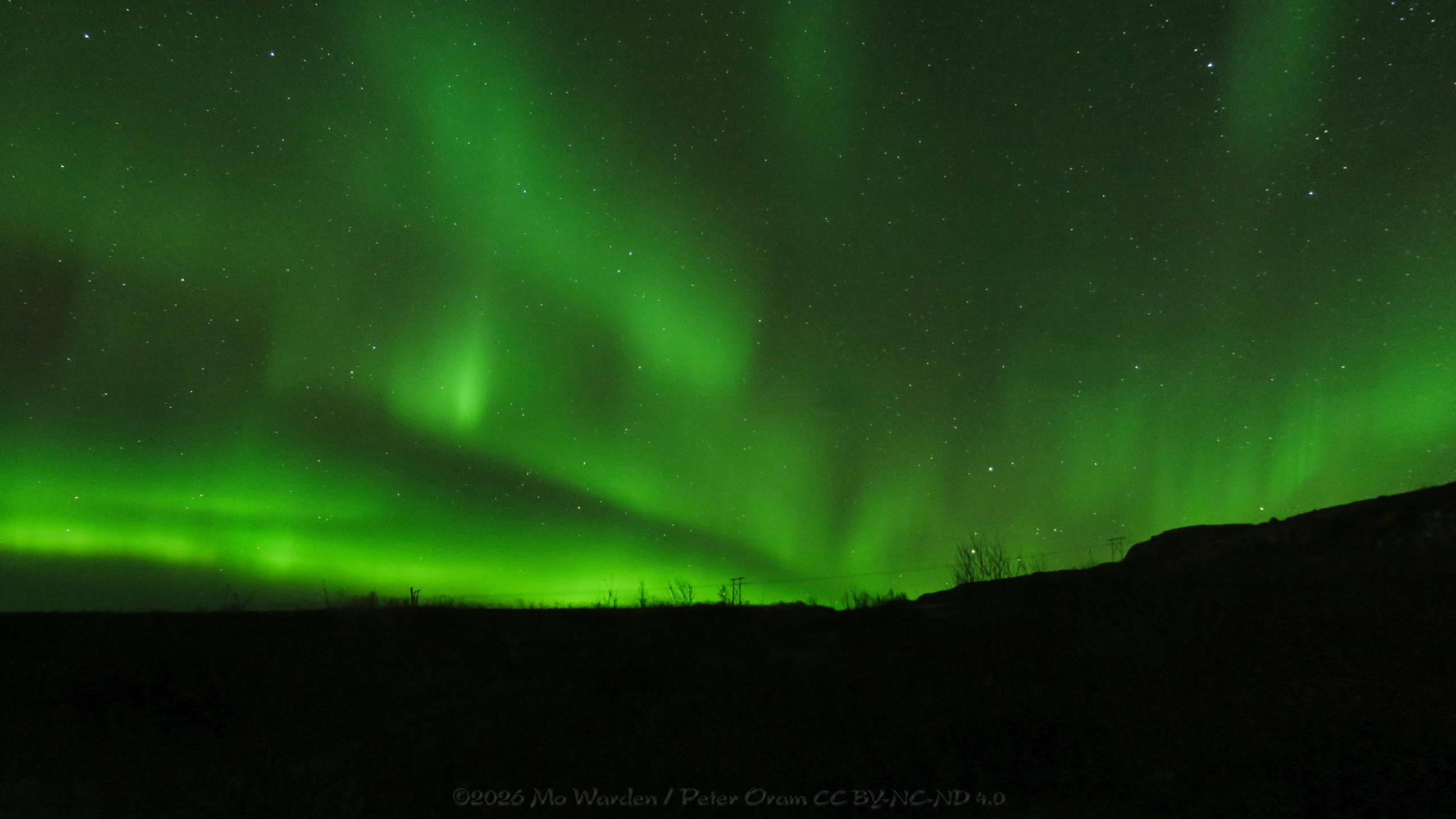 A colour photo of a night sky full of stars. The foreground is in total darkness; a silhouette of a hillside with power pylons and trees along the top. From the lower centre right of the shot, bright green bands of light are radiating. Some parts are tall standing bars within the ribbons, and some areas are brighter than others. It's a gorgeous auroral display.