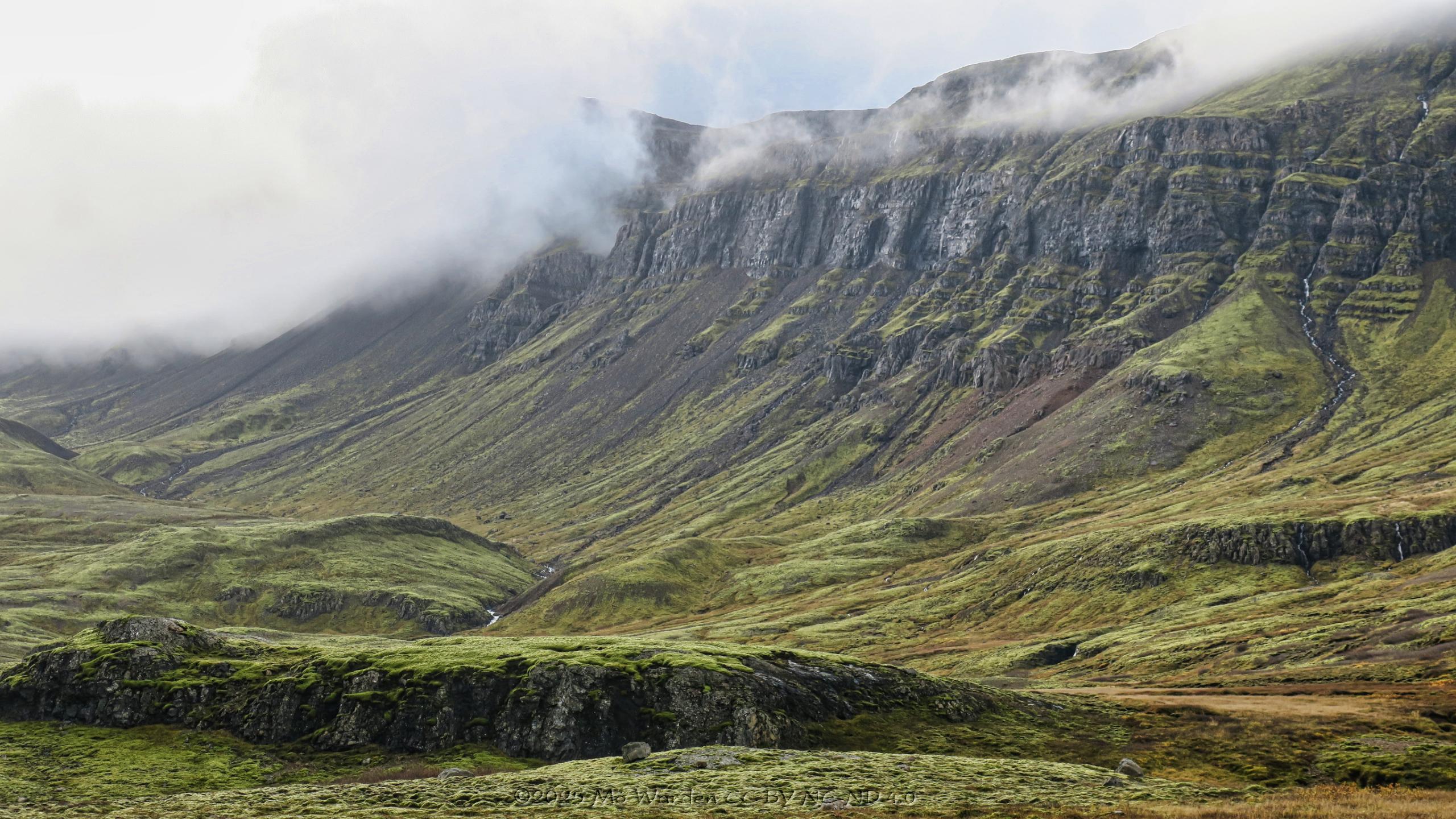 A colour photo of a mountainous landscape. The foreground is mossy green land rising slowly in a glacial valley U-shape to a flatter mesa of bare grey rock. On the right of the shot the grey flanks are topped by scraps of cloud but are still visible. On the left the cloud is denser and the peaks are obscured. Waterfalls and rivulets are dropping in many places, and in the lower foreground are multiple streams of white water.