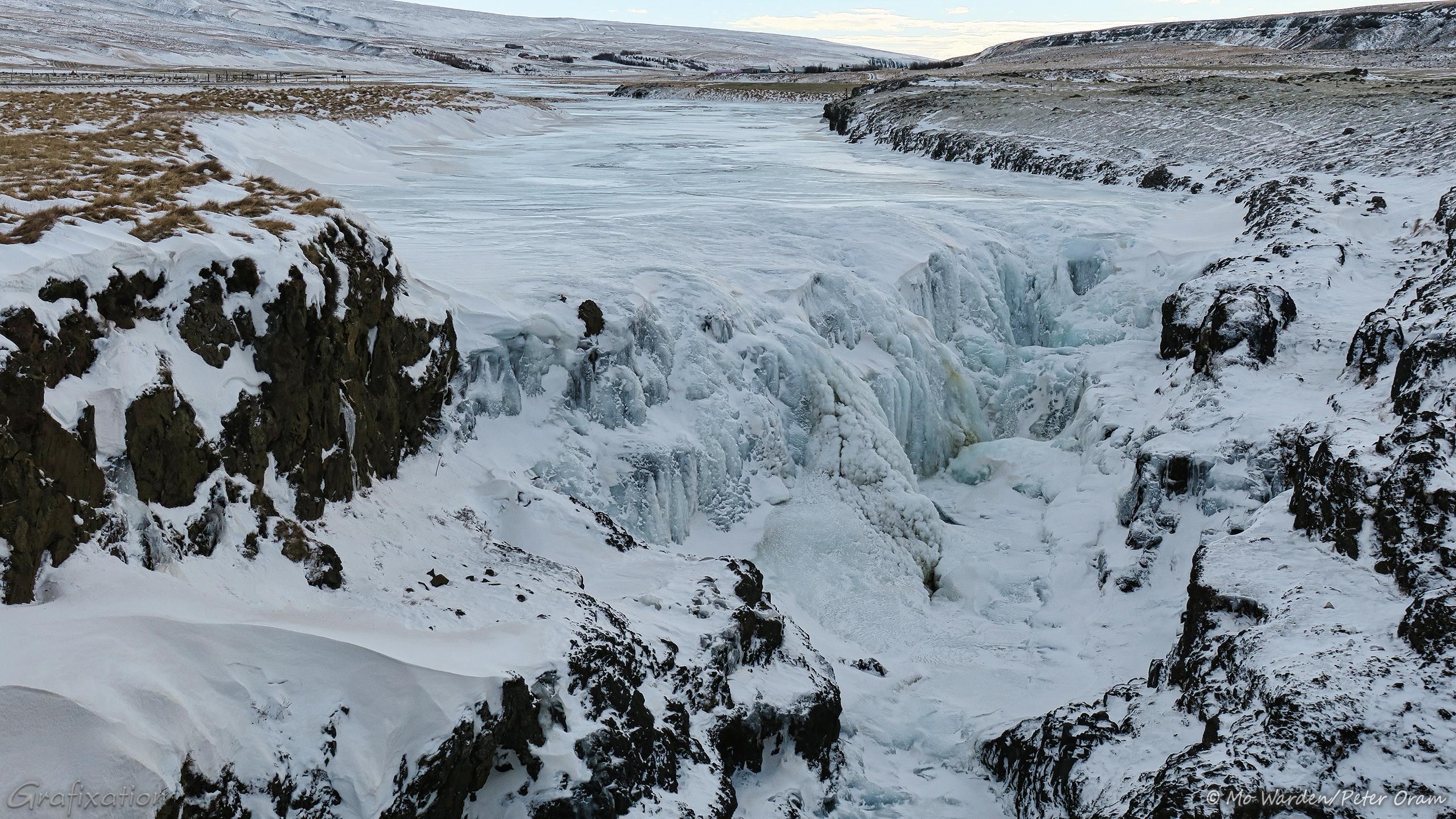 A colour photo of a frozen waterfall in a landscape covered in ice and snow. At the top of the shot in the centre is a river, usually running towards the viewpoint but currently frozen almost stationary. The river banks are quite flat but lead to mountains on either side in the distance. The river drops over a straight crest which is set diagonally in the streambed. From there it drops over rocks which narrow into a gulley, which is completely covered in ice. The rocks on either side of the viewpoint are topped with wind-sculpted snow drifts.